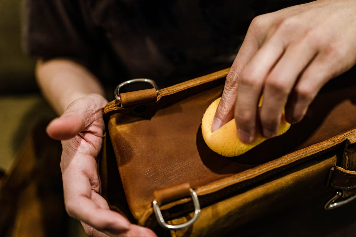 Closeup hand with yellow soft sponge cleaning stains on the surface of the vintage brown bag.